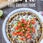 Taco pie filled with ground beef, corn, beans, peppers, covered with cheese tomatoes and green onions in a pie shell. Cutting board with lime and tomatoes in the background