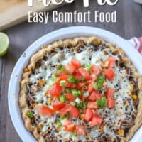 Taco pie filled with ground beef, corn, beans, peppers, covered with cheese tomatoes and green onions in a pie shell. Cutting board with lime and tomatoes in the background