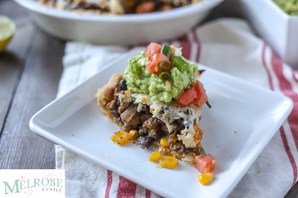Slice of taco pie with ground beef, beans, pepper, and corn with cheese and baked in a pie crust and topped with guacamole and tomatoes on a white plate and a tan an red napkin.