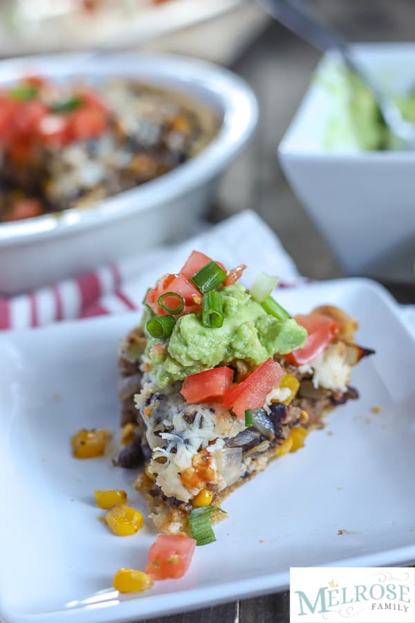 Slice of taco pie with ground beef, beans, pepper, and corn with cheese and baked in a pie crust and topped with guacamole and tomatoes on a white plate and a tan an red napkin with taco pie in the and bowl of guacamole in the background.
