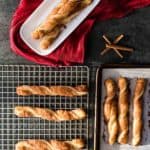 cinnamon twists cooling on a baking rack with more cinnamon twists to the right one a cookie sheet and some in a white dish behind it