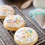 3 sugar cookies with buttercream frosting with sprinkles on a wire rack with a napkin and bowl of frosting behind