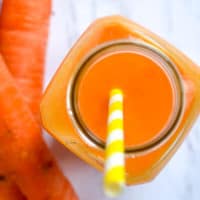 orange carrot smoothie with a yellow straw in a glass bottle with carrots to the left