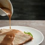 Gravy in a gravy boat being poured over sliced turkey on a plate