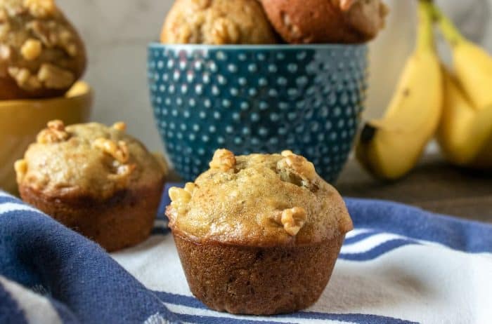 Golden banana nut muffins, looking super filling with their full, round sides, are nestled in a bowl. The crunchy nuts on top add a tasty contrast, making you yearn to pick one up for a satisfying bite.