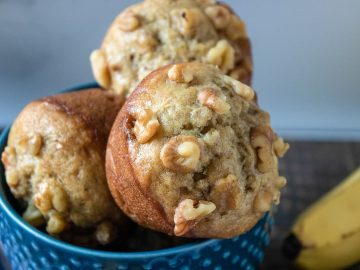 Golden banana nut muffins, looking super filling with their full, round sides, are nestled in a bowl. The crunchy nuts on top add a tasty contrast, making you yearn to pick one up for a satisfying bite.