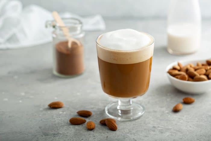 coffee topped with almond milk froth with fresh almonds scattered on the table and a coffee container on the side
