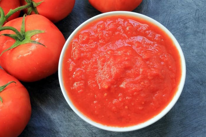 close up of bowl with tomato puree and some tomatoes next to it on gray table