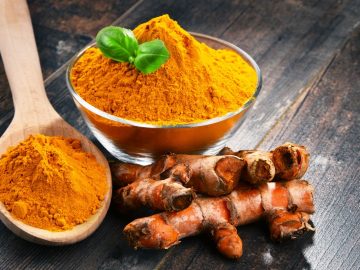 a small glass bowl and wooden spoon full of turmeric powder with fresh turmeric on the sides that are placed on a dark wooden table