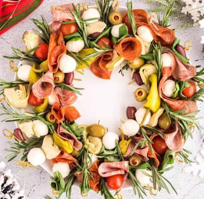 This wreath-shaped board has a stunning arrangement of Italian meats, cheeses, basil, olives, cherry tomatoes, and artichoke hearts on skewers with fresh rosemary sprigs tucked in, creating an unforgettably festive feel.