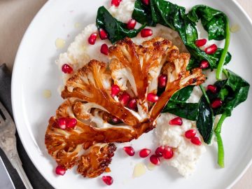 Perfectly roasted cauliflower steak with a glossy, sticky honey sriracha glaze boasting a blend of sweet and spicy flavors. It’s beautifully plated with vibrant pomegranate seeds and lush green spinach, all resting on a smooth bed of mashed cauliflower.