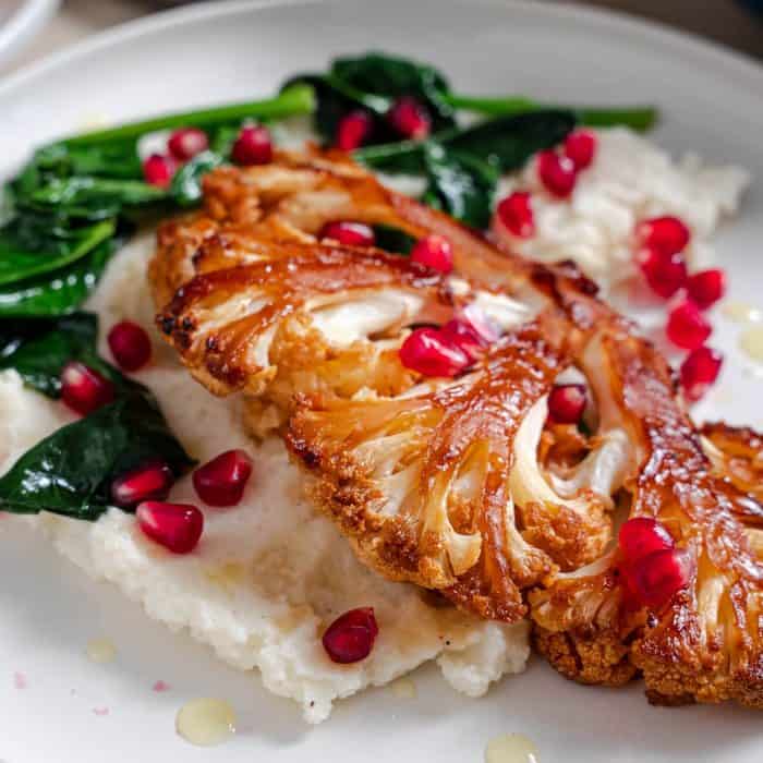 Perfectly roasted cauliflower steak with a glossy, sticky honey sriracha glaze boasting a blend of sweet and spicy flavors. It’s beautifully plated with vibrant pomegranate seeds and lush green spinach, all resting on a smooth bed of mashed cauliflower.
