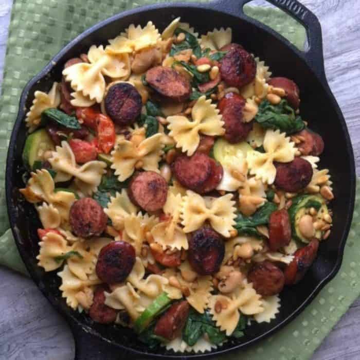 a photo of smoked sausage white bean and spinach pasta with toasted pine nuts placed on a black pan