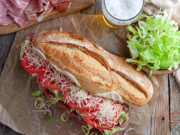 a photo of toasted italian sandwich placed on a wooden table with veggies on the side