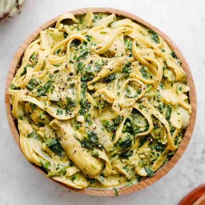 a photo of vegan spinach artichoke pasta on a round wooden plate