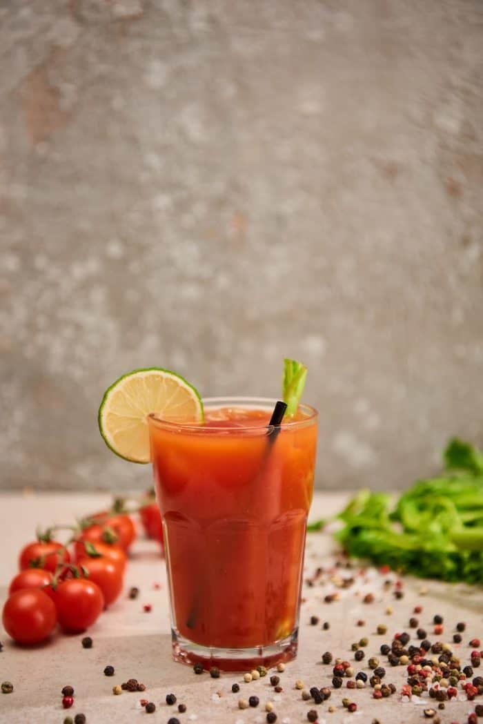 Selective focus of bloody mary cocktail in glass with straw and lime near salt, pepper, tomatoes and celery on grey background