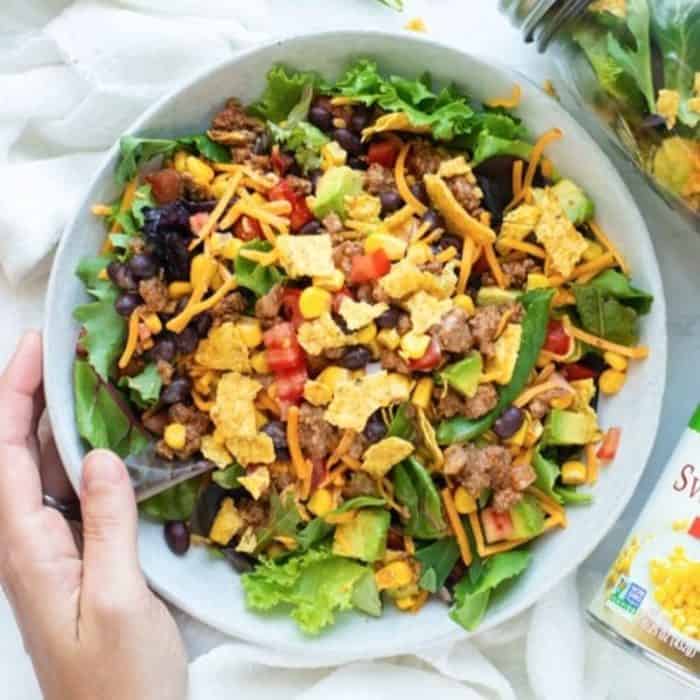 a woman's hand holding a white bowl filled with taco salad