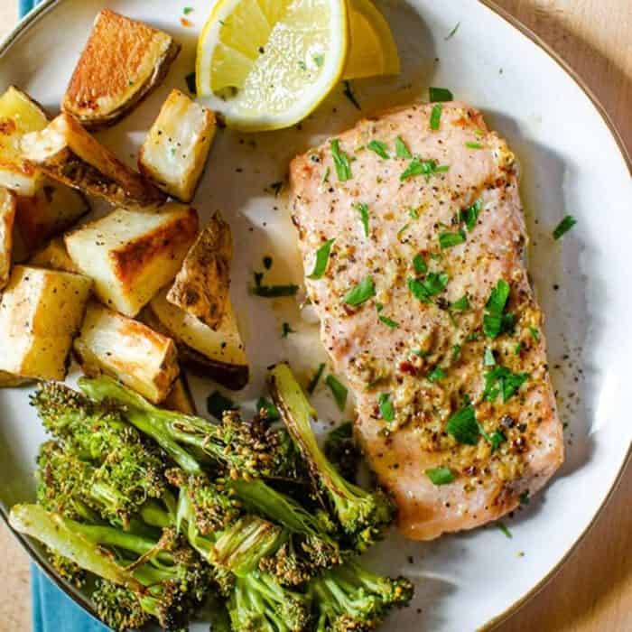 a photo of a white plate filled with baked trouts fillets and roasted veggies and slice of lemon on the side