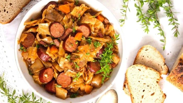 a photo of crockpot cabbage sausages on a white casserole with slice of breads on the side