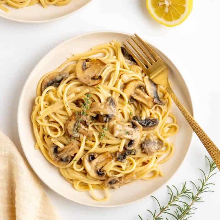 a photo of creamy Vegan Mushroom Pasta on a white round plate with gold fork  on the side