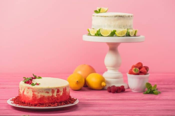 Cake decorated with currants and mint leaves near fruits and white cake on pink wooden surface isolated on pink