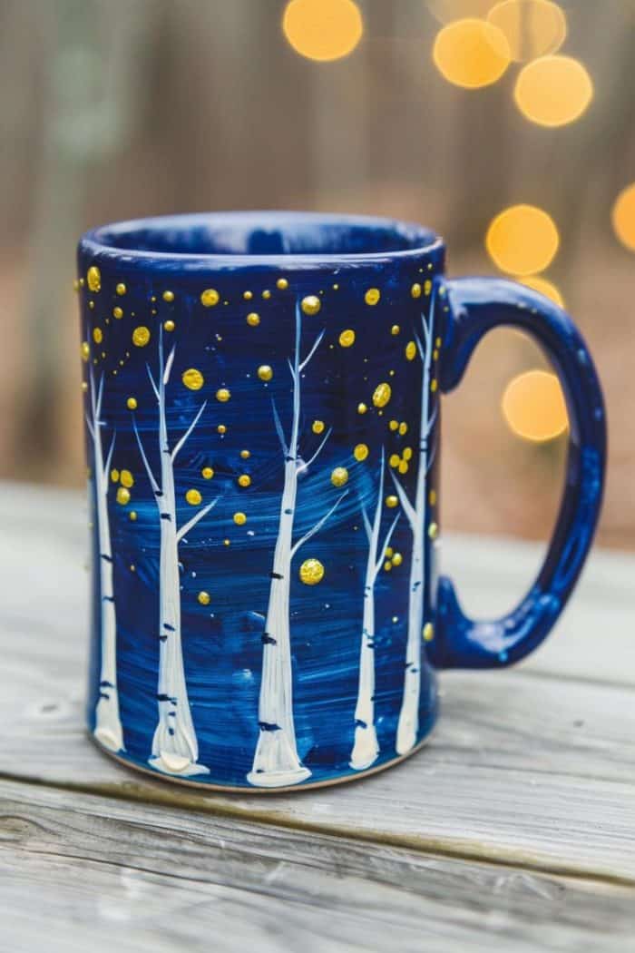 The mug is transformed into an enchanted forest, with bare white trees amidst gold stars. The background is made with a mix of blue and white paint to create depth and mimic the look of a mystical night.
