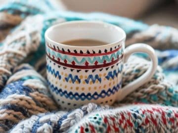 The surface of this mug is painted to resemble the comforting texture of a knitted sweater, complete with interlocking stitches and a pattern that invites one to wrap their hands around it as if warming up by the fire. 