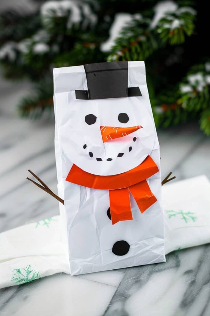 The happy snowman has a carrot nose and a scarf cut out of orange paper and eyes, buttons, and a hat cut out from black paper, creating a friendly face on the white paper bag base.