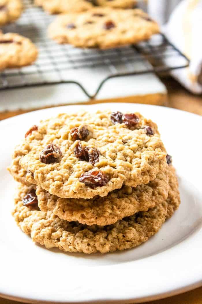 three pieces of freshly cooked oatmeal raisin cookies on a white plate with other cookies placed on a cooling rack on the background