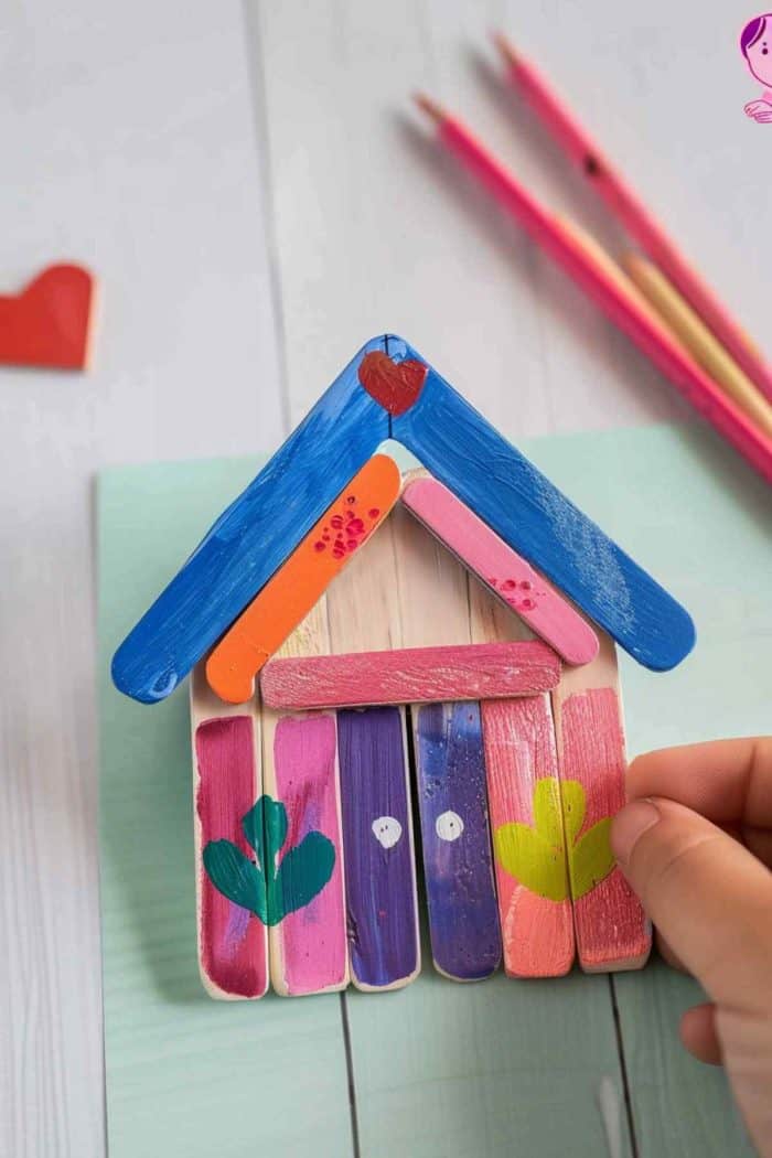 On a piece of colored paper, attach popsicle sticks that form a house shape. Cut two large sticks and use smaller sticks to create a triangular roof with a dynamic look, then use five sticks to create the rest of the house. 