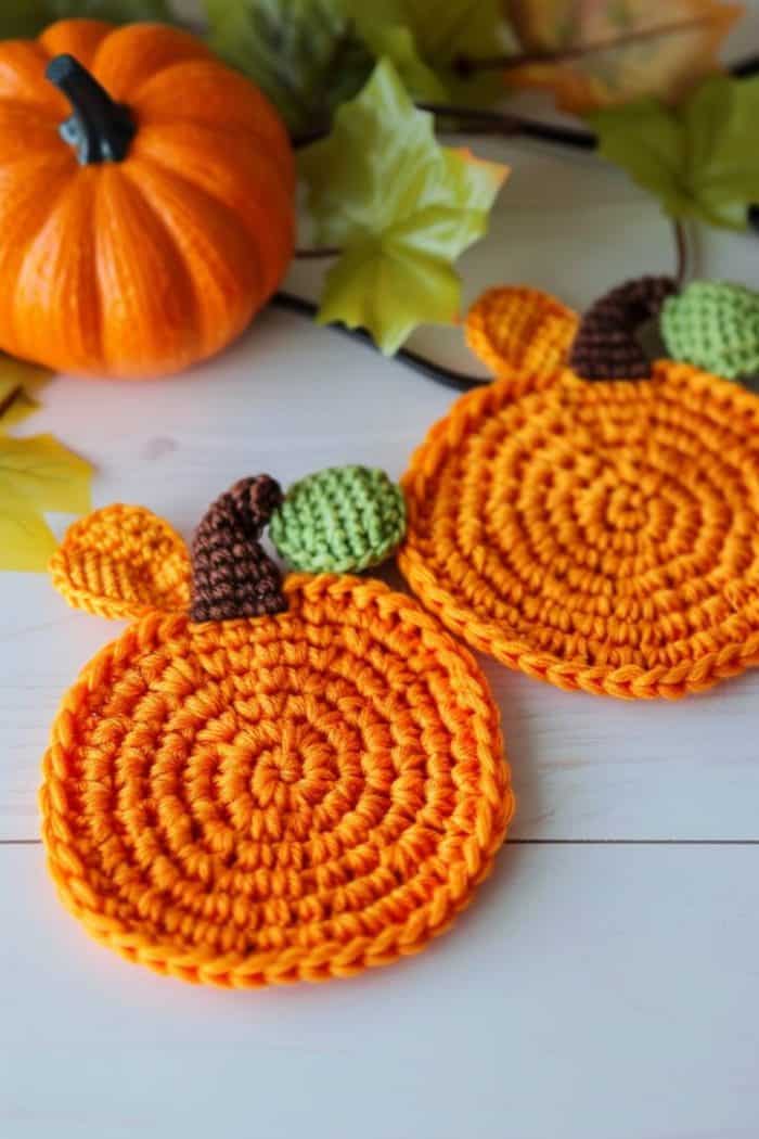 Crochet small, round pumpkin coasters using orange yarn and add a brown crocheted stem and crocheted leaves in different colors on top.