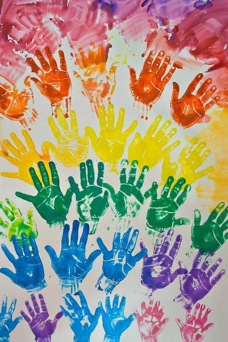 Children press their paint-coated hands onto a canvas, forming a colorful array of handprints that bloom into a personal rainbow. 