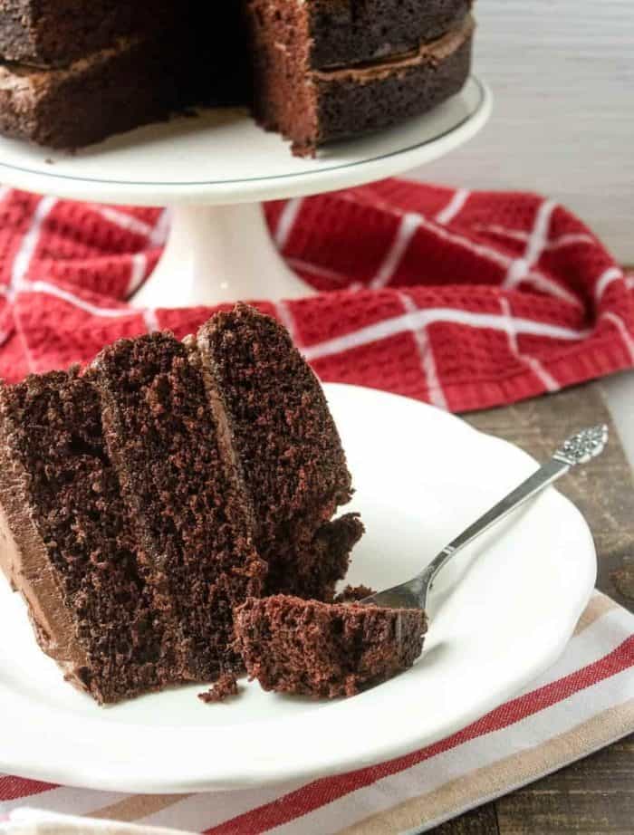 A slice of decadent chocolate layer cake with three moist, rich layers boasting a tender crumb. Smooth, glossy chocolate frosting sweeps in between each layer and across the top of the cake, creating a heavenly dessert.