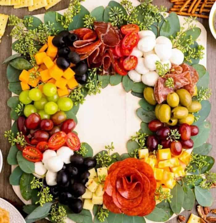 Christmas wreath charcuterie board idea with a variety of cheeses, meats, tomatoes, olives, and fruits on verdant eucalyptus leaves.