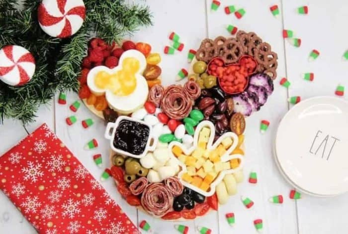 Mickey Mouse-themed Christmas charcuterie board made of olives, tomatoes, raspberries, pretzels, and candies, the board features Mickey Mouse Goldfish crackers, cheese cubes in a Mickey Mouse hand, and savory salami rolled up into a Mickey Mouse head