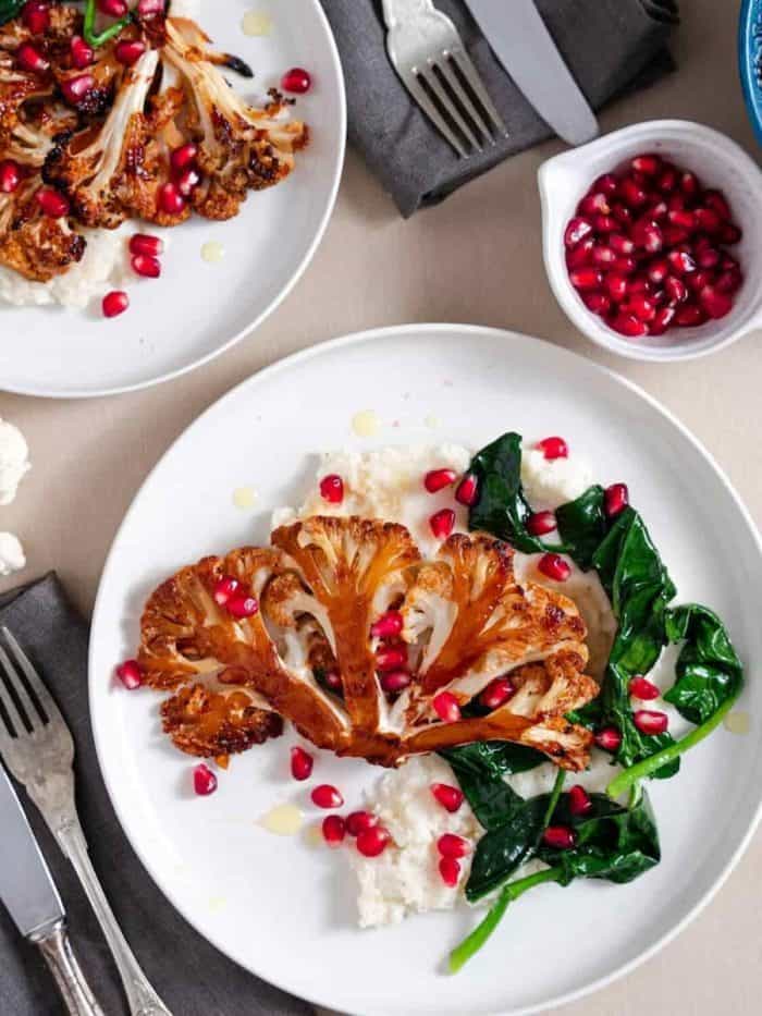 Perfectly roasted cauliflower steak with a glossy, sticky honey sriracha glaze boasting a blend of sweet and spicy flavors. It’s beautifully plated with vibrant pomegranate seeds and lush green spinach, all resting on a smooth bed of mashed cauliflower.