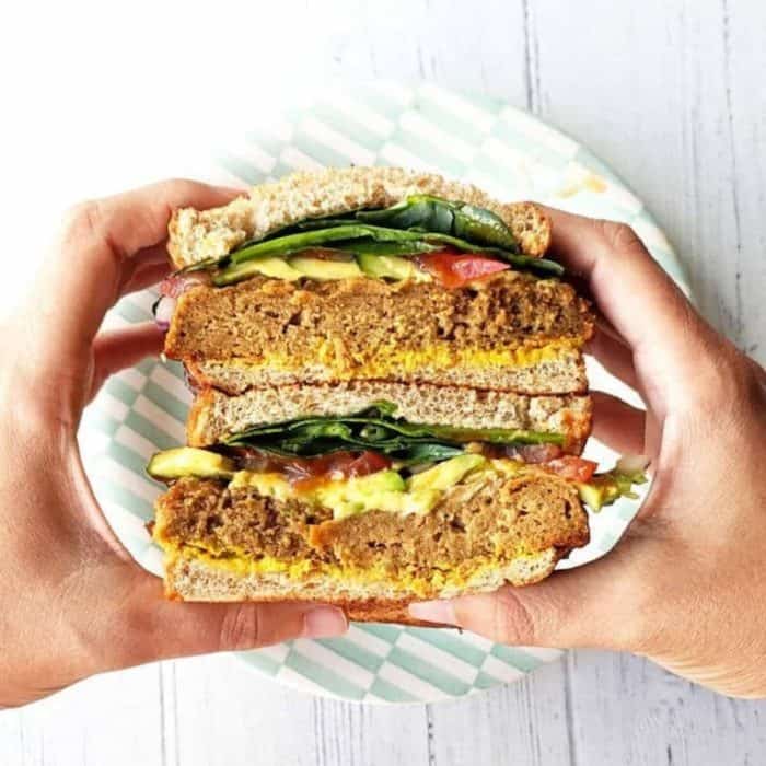 a photo of a hands holding a whole bbg seitan vegan sandwich on a white wooden surface background