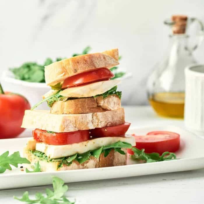 layers of caprese vegan sandwich on a white rectangular plate with tomato and green leaves on side