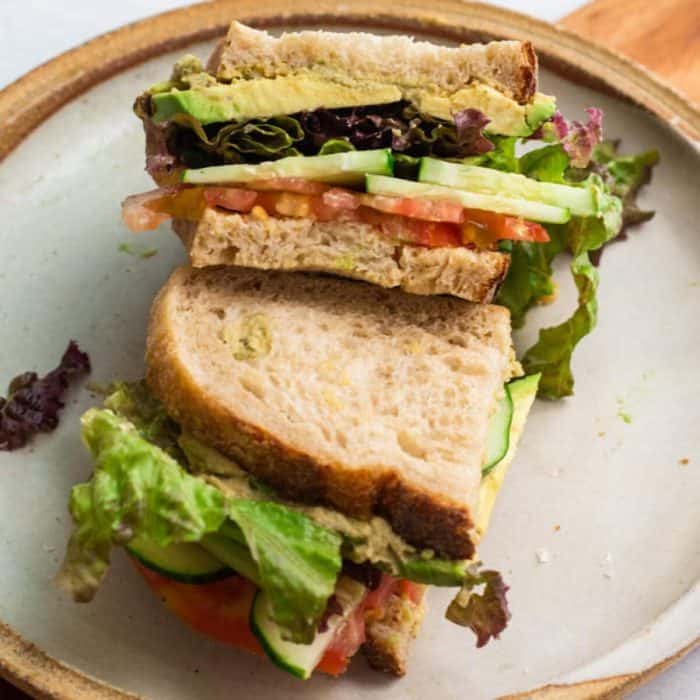 a close up photo of two layers of cucumber hummus loaded with cucumber sandwich on top of a wooden chopping board