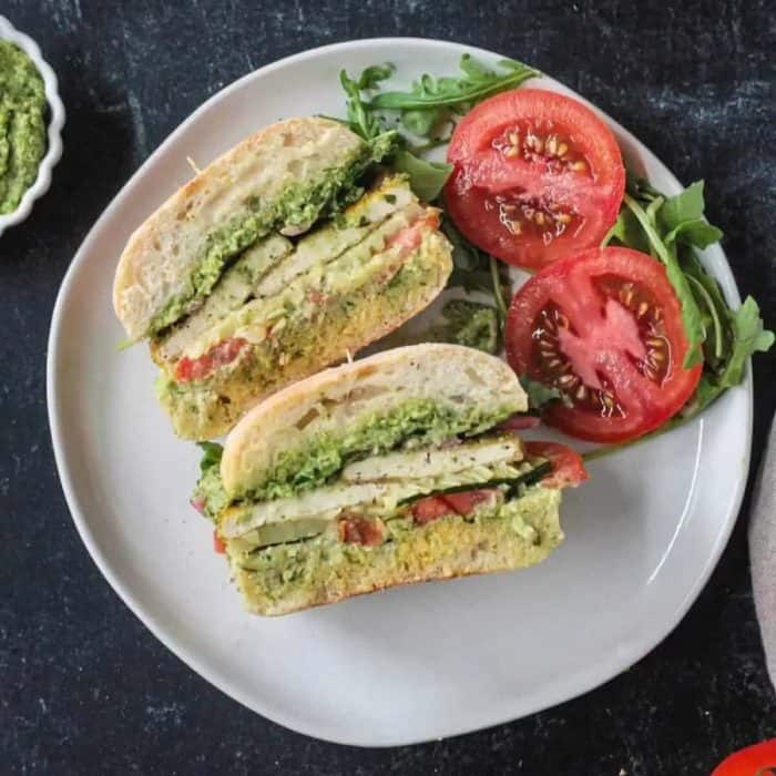 a flatlay photo of two slices of pesto tofu vegan sandwich on a white plate with tomato and lettuce on sides