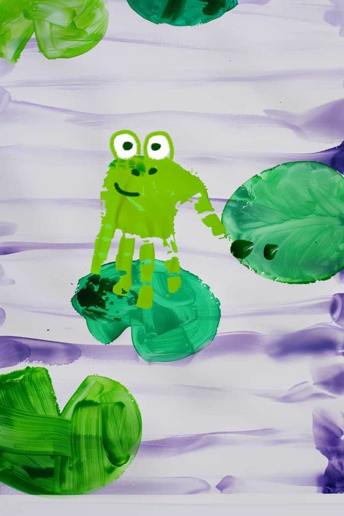 Form a pond scene with green handprints for lily pads, adding small handprint frogs sitting atop. Use lighter green paint for the frogs and darker for the pads, and add details like eyes and ripples in the water to complete the tranquil setting.