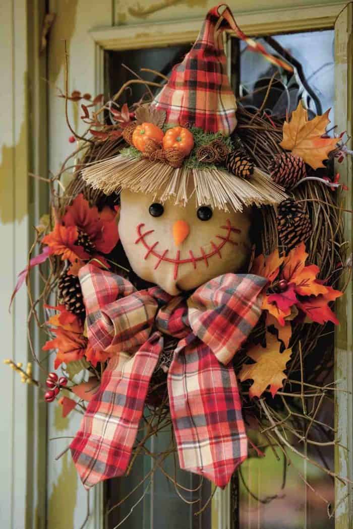 A wreath featuring a scarecrow face with a plaid hat and bow, surrounded by autumn leaves and pinecones, hangs on a door.