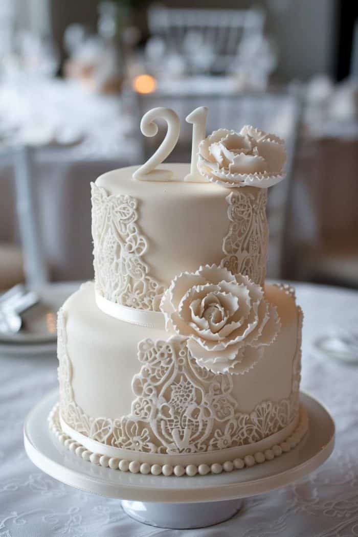 A two-tiered white cake adorned with intricate lace-like icing patterns, topped with the number 21 and two large, detailed sugar roses. The cake sits on a white tablecloth with blurred dining elements in the background.