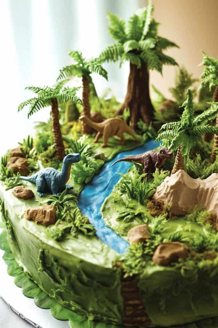 A cake decorated with green icing, small trees, and dinosaur figurines resembling a prehistoric landscape with a river flowing through the center.