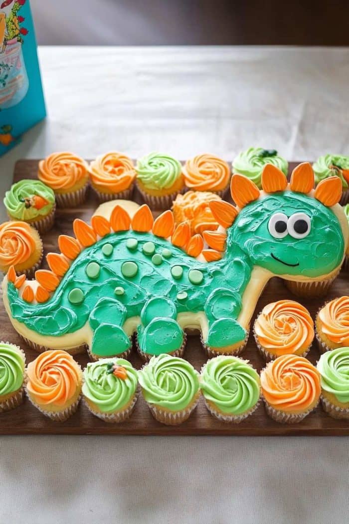 A dinosaur-themed cupcake cake with a green and orange dinosaur-shaped cake centerpiece, surrounded by green and orange frosted cupcakes on a wooden board.