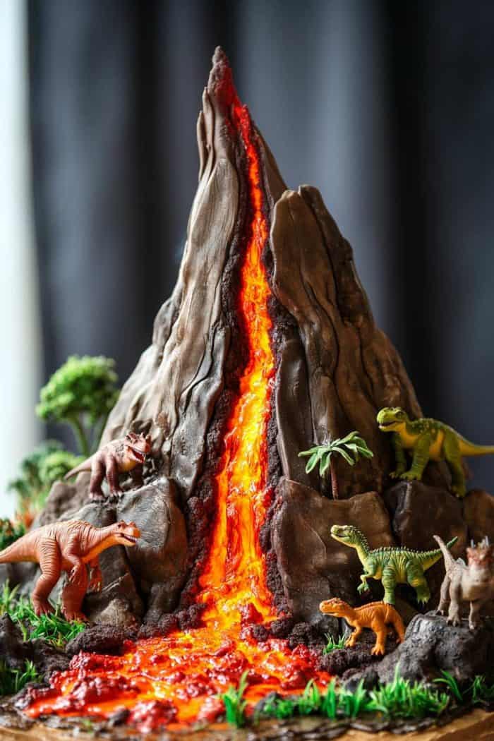 A detailed model of a volcano cake with flowing lava, surrounded by various dinosaur figurines including T-Rex and triceratops, set against a dark background.