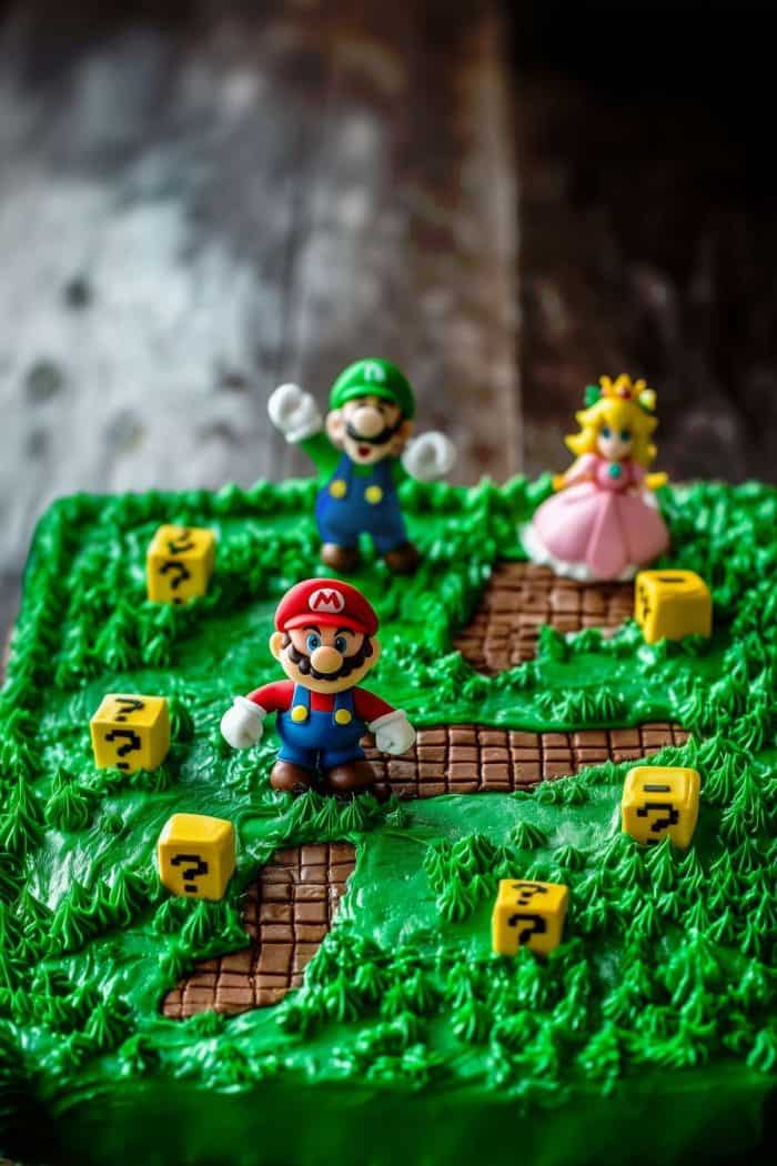 A cake topped with green icing resembling grass, arranged with small figurines of characters from the Mario series and yellow question mark blocks, styled to look like a game scene.