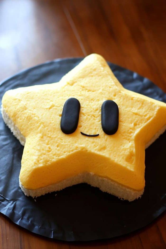 A star-shaped cake with a yellow top and a white base, featuring two black fondant eyes and a small smiling mouth. The cake is placed on a dark surface.