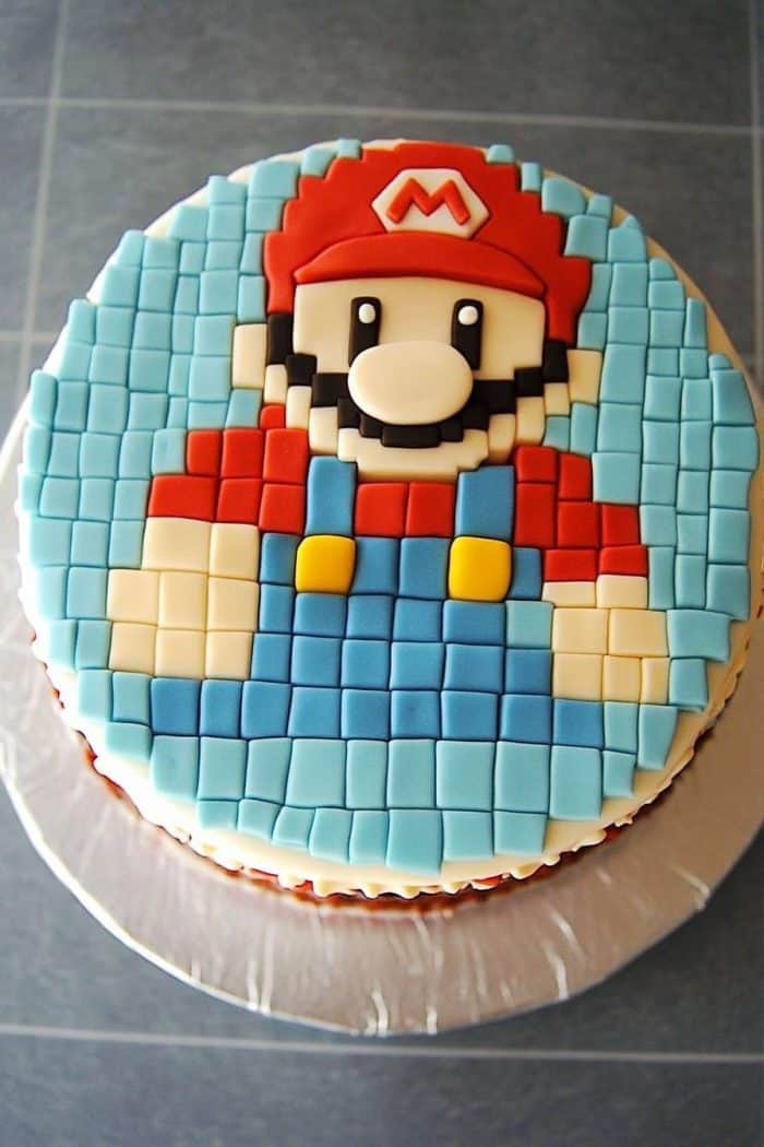 A round cake with a pixelated design of a character wearing a red hat, red shirt, and blue overalls made from icing. The background is blue and the cake sits on a white cake board.