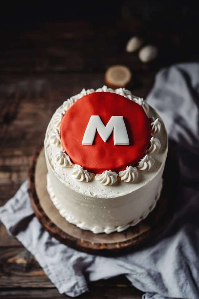A white frosted cake topped with a red fondant featuring a large white letter "M" in the center, surrounded by white icing decorations, placed on a wooden board.